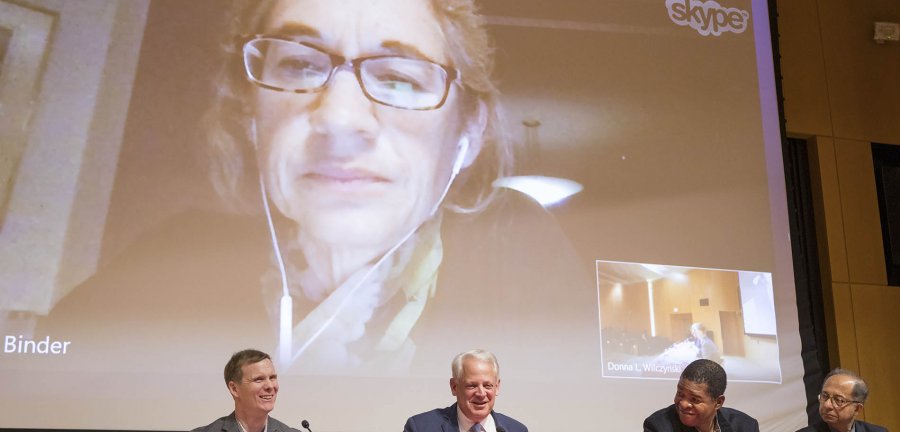 Four men sitting in front of a screen with a woman joining virtually.