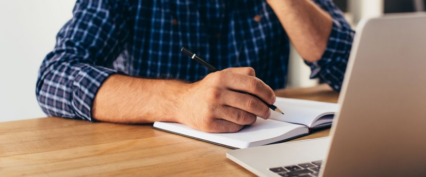 Arm of person taking notes in pad in front of an open laptop.