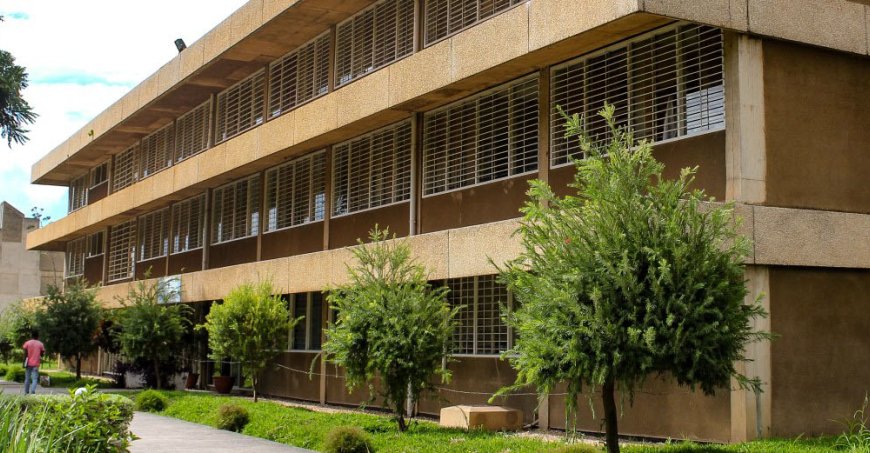Three-story tan concrete building with trees in front and additional building in the background.