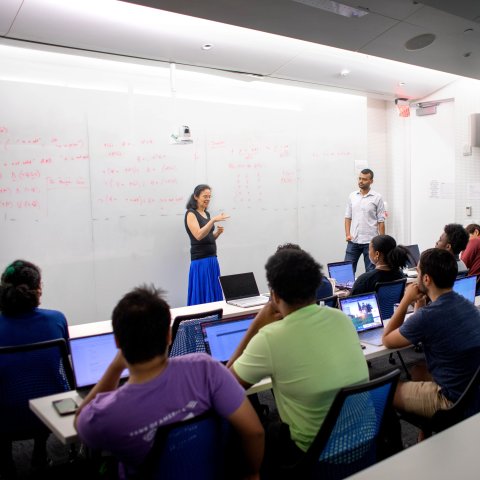 Faculty member standing in front of dry erase board with rows of seated students. 