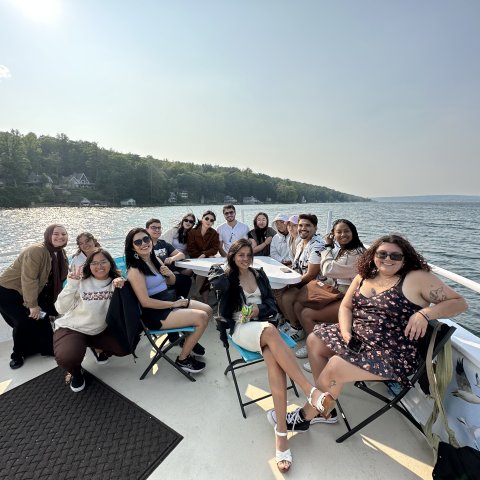 A group of students sit together at the front of a boat on Cayuga Lake, smiling for a photo. 