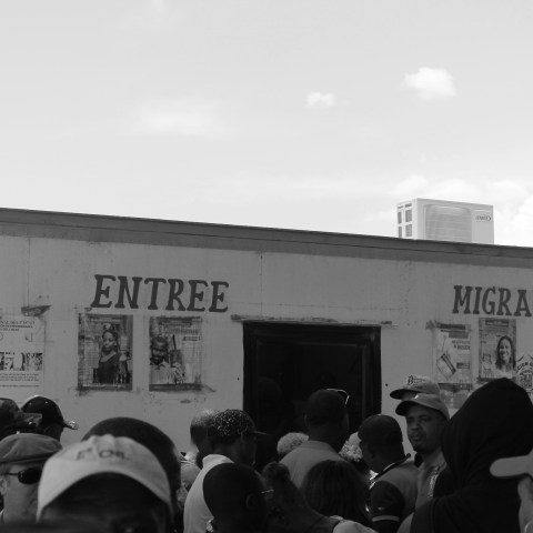 In black and white: a crowd of people stand in front of a building that reads "Entree Migracion."