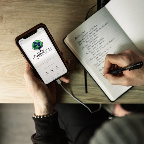 A person takes notes in a notebook while listening to the Migrations: A World on the Move podcast.