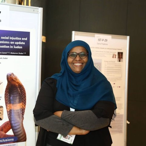 Rania Mohamed Hassan Baleela standing in front of a poster displaying her work. 